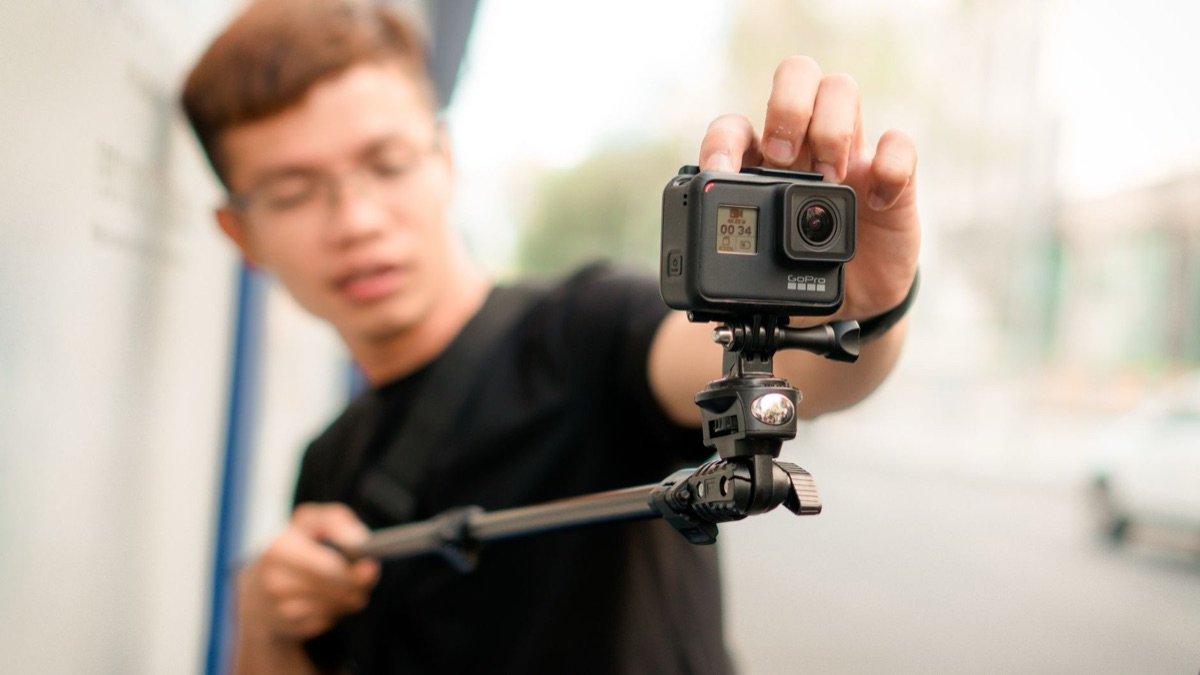 A person's hands holding a compact action camera while walking through a forest.